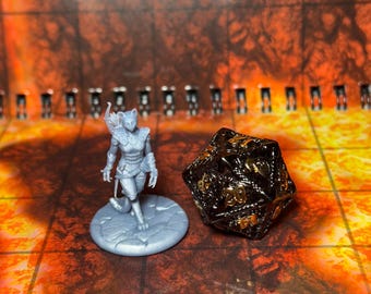 May include: A gray plastic miniature figurine of a cat-like humanoid with wings, wearing armor and holding a sword, standing on a gray base. A black and gold metal twenty-sided die is next to the figurine.