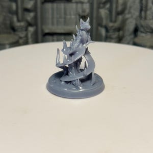 May include: A gray miniature figurine of a fantasy creature with a detailed design. The figure has a circular base and a complex, spiraling form. The background includes a blurred image of a tavern sign and a stone wall.