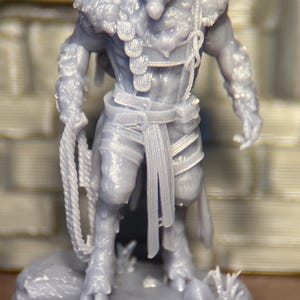 May include: A gray figurine of a goat-headed humanoid creature, possibly for tabletop gaming. The figure has horns, fur, and is holding a rope. It is standing on a circular base with some grass.