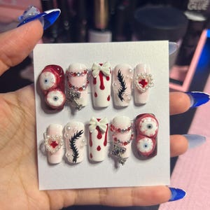 May include: A set of ten artificial nails with various designs. The nails feature a mix of white, red, and clear colors, adorned with embellishments like bows, rhinestones, and eye-shaped designs. Some nails have a dripping blood effect, while others have a feather design.