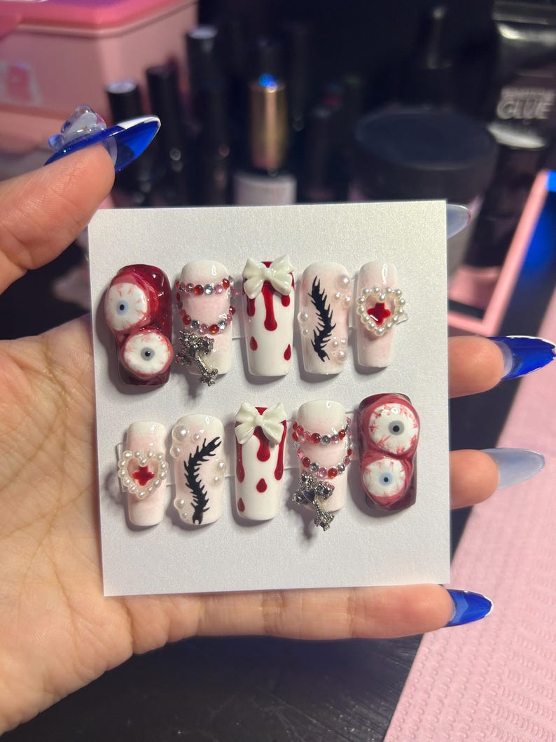 May include: A set of ten artificial nails with various designs. The nails feature a mix of white, red, and clear colors, adorned with embellishments like bows, rhinestones, and eye-shaped designs. Some nails have a dripping blood effect, while others have a feather design.