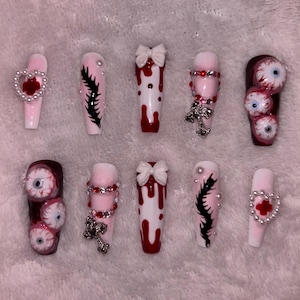 May include: A set of ten long, coffin-shaped artificial nails. Designs include dripping blood, bows, cross charms, and eyeball accents. Colors are primarily pink, red, white, and black. Some nails feature heart-shaped embellishments.