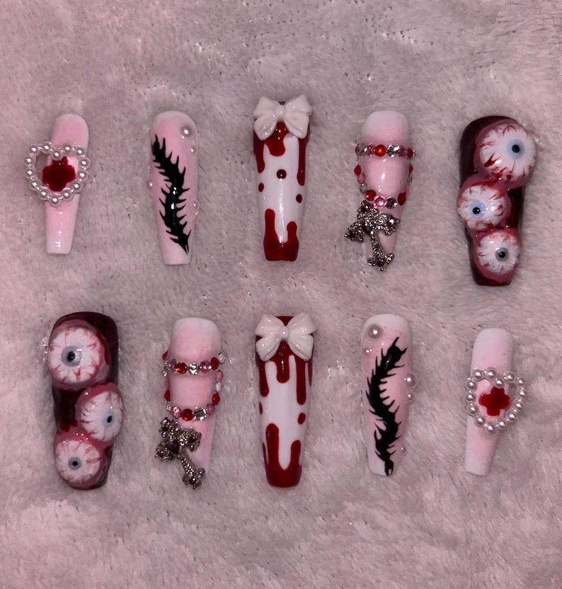 May include: A set of ten long, coffin-shaped artificial nails. Designs include dripping blood, bows, cross charms, and eyeball accents. Colors are primarily pink, red, white, and black. Some nails feature heart-shaped embellishments.