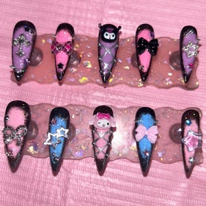 May include: A set of ten long, pointed artificial nails. Each nail features a unique design with glitter, bows, stars, and character accents in pink, purple, and blue. Some nails have silver studs.