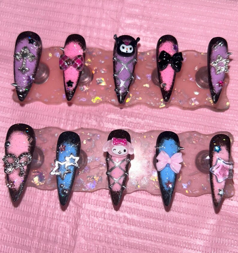 May include: A set of ten long, pointed artificial nails. Each nail features a unique design with glitter, bows, stars, and character accents in pink, purple, and blue. Some nails have silver studs.