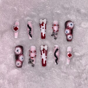 May include: A set of ten coffin-shaped press-on nails with various designs. Some nails feature dripping blood, bows, and cross charms. Others have eyeball designs, black feather-like art, and heart accents. The nails are primarily white, red, and pink.