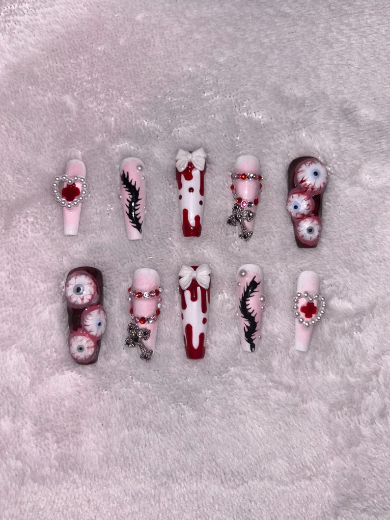 May include: A set of ten coffin-shaped press-on nails with various designs. Some nails feature dripping blood, bows, and cross charms. Others have eyeball designs, black feather-like art, and heart accents. The nails are primarily white, red, and pink.