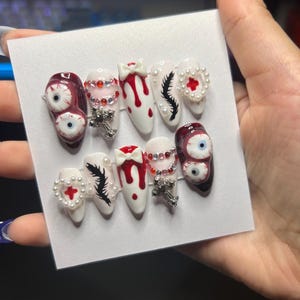 May include: A set of press-on nails with a spooky theme. The nails feature designs like blood drips, eyeballs, crosses, and feather accents. Some nails are adorned with rhinestones and pearl accents, creating a unique and eye-catching look.