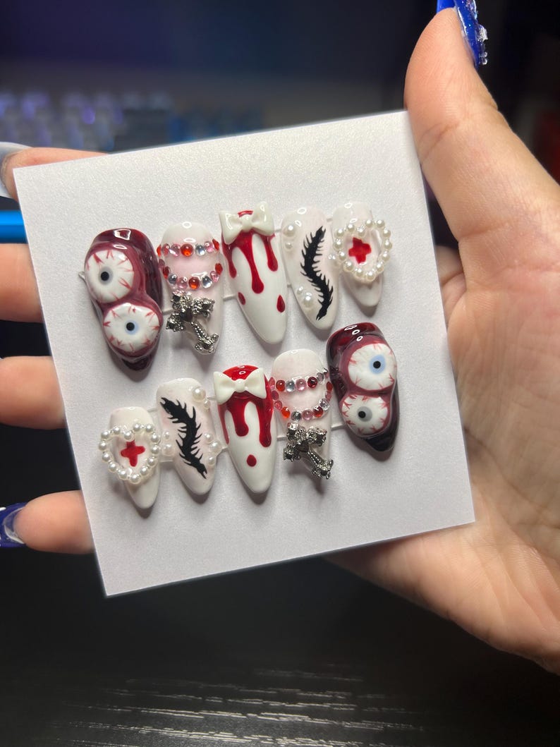 May include: A set of press-on nails with a spooky theme. The nails feature designs like blood drips, eyeballs, crosses, and feather accents. Some nails are adorned with rhinestones and pearl accents, creating a unique and eye-catching look.