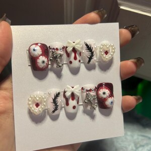 May include: A set of press-on nails with a spooky theme. Designs include red and white with blood drop accents, eyeball art, black feather details, pearl heart accents, and silver cross charms. The nails are held on a white card.