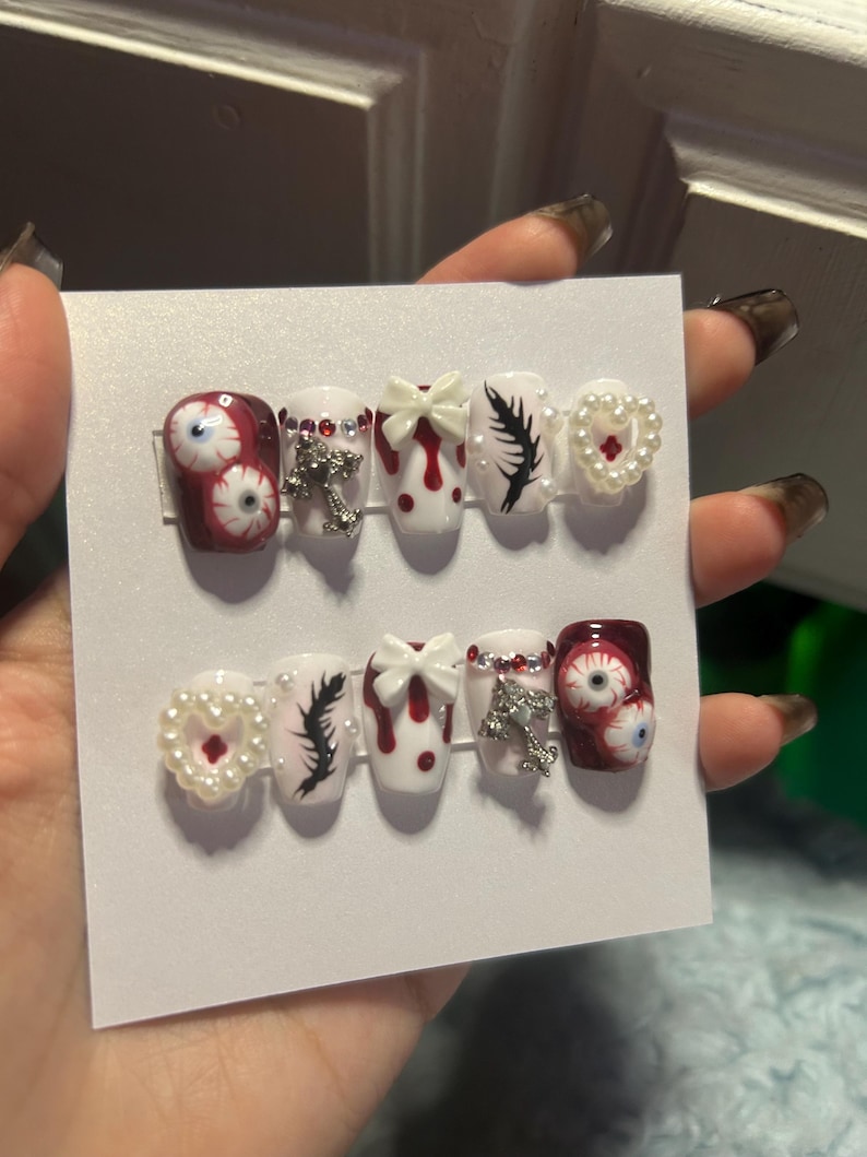 May include: A set of press-on nails with a spooky theme. Designs include red and white with blood drop accents, eyeball art, black feather details, pearl heart accents, and silver cross charms. The nails are held on a white card.