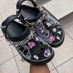 Op de afbeelding: Zwarte Crocs klompen met een punkrockthema. De schoenen zijn bedekt met strass, bedels en spikes. De bedels omvatten een cartoonfiguur, een auto en een hart. De schoenen hebben een zilveren ketting eromheen.