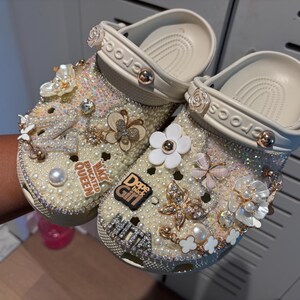 Op de afbeelding: Een paar witte klompen versierd met parels, strass en bedels. De bedels omvatten een vlinder, een bloem en een bedel met de tekst "Dope Black Girl".