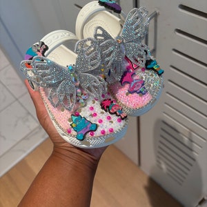 May include: A pair of white clogs decorated with large silver rhinestone butterfly charms, pink and white pearls, and colorful cartoon character charms. The shoes feature a pivoting heel strap and a perforated upper surface covered in crystals.
