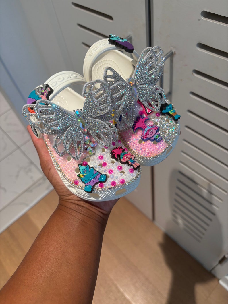 May include: A pair of white clogs decorated with large silver rhinestone butterfly charms, pink and white pearls, and colorful cartoon character charms. The shoes feature a pivoting heel strap and a perforated upper surface covered in crystals.