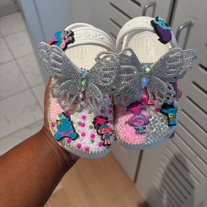 May include: A pair of white clogs decorated with silver butterfly charms, pink and white pearls, and colorful cartoon character charms. The surface of the clogs is covered in small clear rhinestones, and the heel straps feature additional character charms.