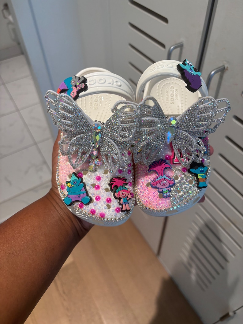 May include: A pair of white clogs decorated with silver butterfly charms, pink and white pearls, and colorful cartoon character charms. The surface of the clogs is covered in small clear rhinestones, and the heel straps feature additional character charms.