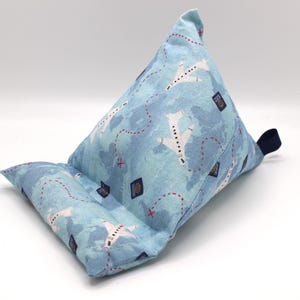 May include: A blue and white fabric tablet pillow with a travel-themed pattern. The pillow features a light blue background with white airplanes, red dotted lines, and passport stamps. A dark blue fabric loop is attached to the side.