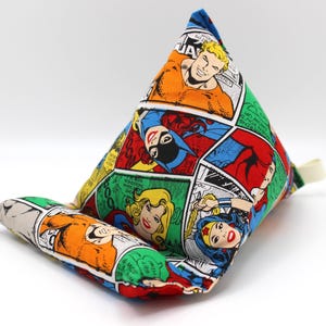 May include: A comic book-themed tablet stand featuring panels with colorful illustrations of superheroes. The stand is triangular with a supportive base and a small fabric loop. The fabric is a mix of red, blue, green, and yellow.
