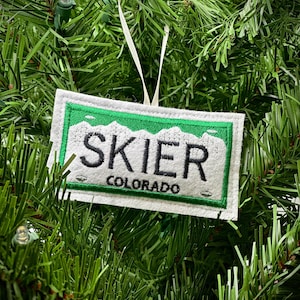 Könnte beinhalten: Ein weiß-grüner, gestickter Ornament mit dem Wort "SKIER" und "COLORADO" in schwarzer Schrift. Der rechteckige Ornament hat ein weißes Band zum Aufhängen. Der Ornament hängt an einem grünen Weihnachtsbaum.