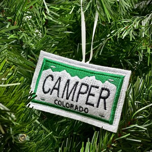 Könnte beinhalten: Ein rechteckiger, weiß-grüner, gestickter Ornament mit dem Wort "CAMPER" und "COLORADO" in Schwarz. Das Ornament hat einen grünen Rand und ein Bergdesign. Ein weißes Band ist zum Aufhängen befestigt.