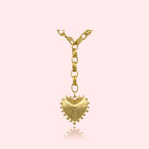 May include: A gold-colored necklace with a heart-shaped pendant. The pendant has a textured surface and a beaded edge. The necklace chain is made of large, linked segments. The background is a soft pink.