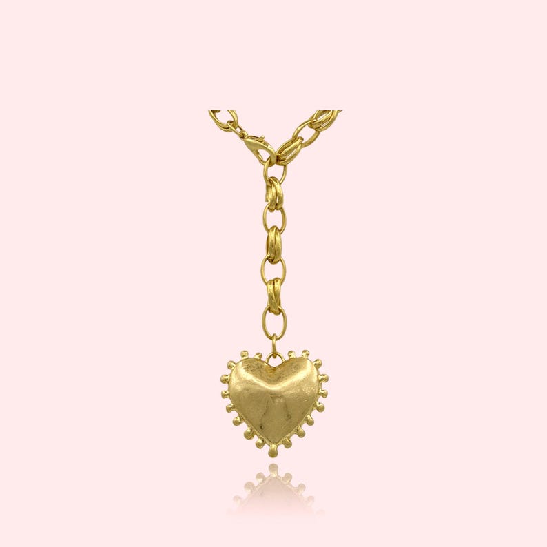 May include: A gold-colored necklace with a heart-shaped pendant. The pendant has a textured surface and a beaded edge. The necklace chain is made of large, linked segments. The background is a soft pink.