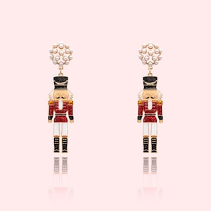 May include: A pair of festive dangle earrings featuring a red, black, and white nutcracker soldier design. Each earring has a gold-colored top with a cluster of small, white pearls. The earrings are set against a soft pink background.