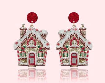 May include: A pair of festive gingerbread house earrings. Each earring features a glittery gold and white house with red and green accents, a red door, and a chimney. The earrings are topped with a red, glittery circle.