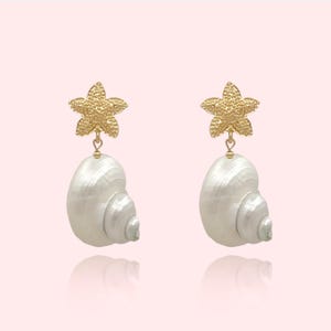 Puede incluir: Un par de pendientes de estrella de mar dorada con colgantes de concha blanca. Las conchas tienen un acabado nacarado y un diseño en espiral.