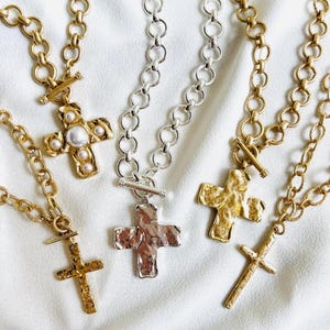 May include: Several gold-toned and silver-toned cross necklaces with varying chain styles and textures. Some crosses have a hammered appearance, while others feature pearl accents. The necklaces are displayed on a white, textured surface.