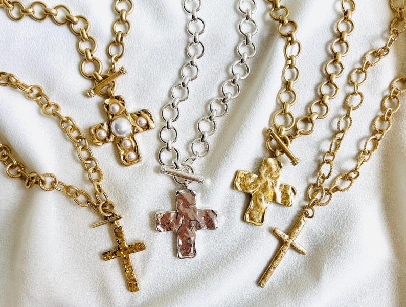 May include: Several gold-toned and silver-toned cross necklaces with varying chain styles and textures. Some crosses have a hammered appearance, while others feature pearl accents. The necklaces are displayed on a white, textured surface.