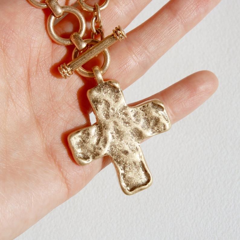May include: A gold-colored necklace with a textured cross pendant. The necklace features a toggle clasp and a chain with circular links. The cross has a hammered appearance, and the metal has a matte finish.