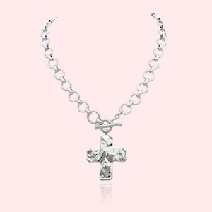 May include: Silver-toned necklace featuring a textured cross pendant. The necklace has a chain of large, round links and a toggle clasp. The cross pendant has a hammered metal appearance, reflecting light. The jewelry is displayed against a soft pink background.
