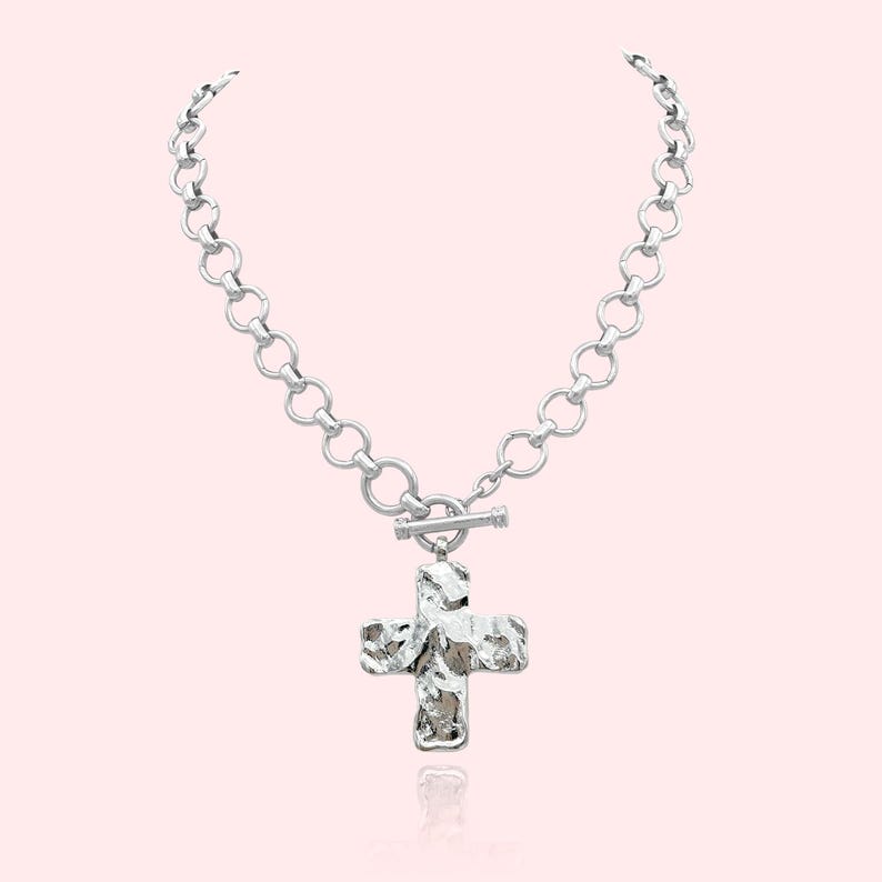 May include: Silver-toned necklace featuring a textured cross pendant. The necklace has a chain of large, round links and a toggle clasp. The cross pendant has a hammered metal appearance, reflecting light. The jewelry is displayed against a soft pink background.