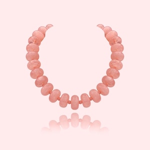 May include: A rose quartz necklace with a soft pink hue. The necklace features a series of rounded, polished beads strung together. The beads have a translucent quality, and the necklace is displayed against a matching pink background.