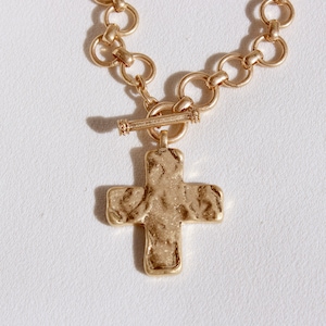 May include: Gold-toned necklace featuring a textured cross pendant. The necklace has a toggle clasp and a chain of interconnected circular links. The cross has a hammered appearance, adding a rustic charm to the jewelry.