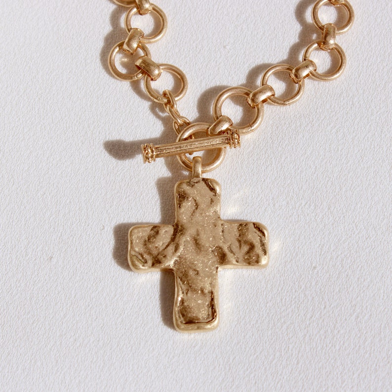 May include: Gold-toned necklace featuring a textured cross pendant. The necklace has a toggle clasp and a chain of interconnected circular links. The cross has a hammered appearance, adding a rustic charm to the jewelry.