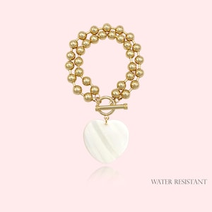 May include: Gold-tone beaded bracelet with a heart-shaped pendant. The bracelet features a toggle clasp and a white, heart-shaped pendant. The words "WATER RESISTANT" are printed at the bottom right of the image.