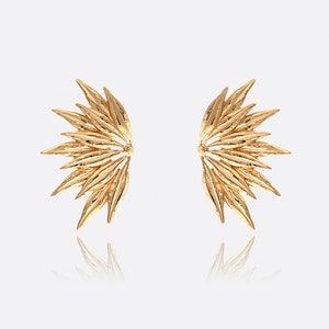 May include: A pair of gold-colored earrings. Each earring is designed in a half-wing shape, with multiple leaf-like structures radiating outwards. The earrings are displayed against a white background, with a reflection visible below.