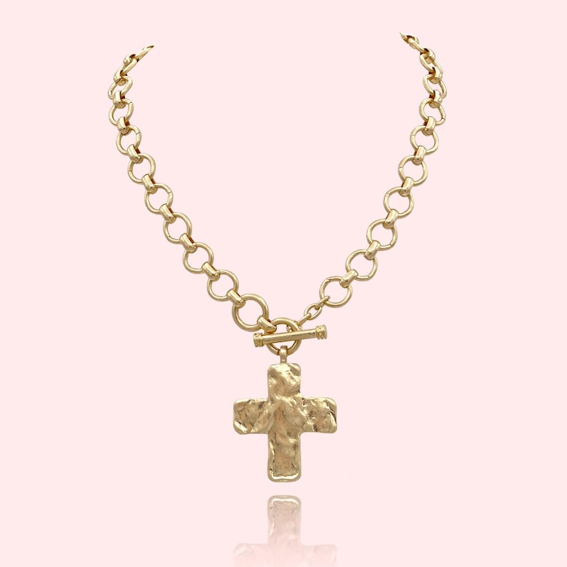 May include: Gold-toned necklace featuring a textured cross pendant. The necklace has a chunky chain design with a toggle clasp. The cross pendant has a rough, hammered appearance. The jewelry is displayed against a light pink background.