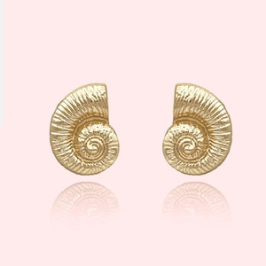 May include: A pair of gold-toned earrings shaped like seashells. The earrings are detailed with a textured surface and a spiral design.