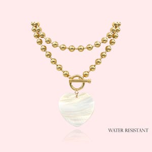 May include: Gold-colored necklace with two strands of small, round beads. A toggle clasp connects the strands. A heart-shaped pendant with a pearlescent finish hangs from the clasp. The text "WATER RESISTANT" is visible.