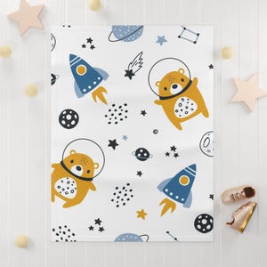 Può includere: Coperta per bambini bianca con un motivo ripetuto di orsi dei cartoni animati in tute spaziali, razzi blu, pianeti, stelle e costellazioni. Gli orsi sono marroni con la pancia bianca. I razzi sono blu con fiammate arancioni.