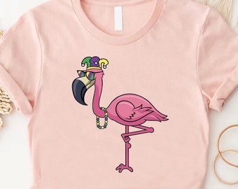 May include: A pink flamingo wearing a purple and green jester hat and sunglasses with a purple and green beaded necklace. The flamingo is standing on one leg and is on a pink t-shirt.