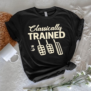 May include: Black t-shirt with the text "Classically TRAINED" in a vintage font, accompanied by illustrations of car pedals. The design is in a cream color. The shirt is folded and knotted at the bottom.