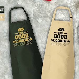 May include: Two aprons with adjustable neck straps and pockets. One apron is green with the text "MR. GOOD LOOKIN' IS COOKIN'" in gold. The other apron is beige with the same text in black.