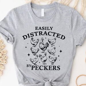 Op de afbeelding: Grijs T-shirt met een zwarte afbeelding die "Gemakkelijk afgeleid door pikken" zegt en een haan en kippen toont.