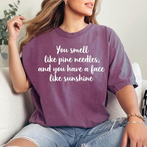 K&ouml;nnte beinhalten: Eine Frau tr&auml;gt ein lila T-Shirt mit dem Text "You smell like pine needles, and you have a face like sunshine" darauf. Sie sitzt auf einem wei&szlig;en Sofa mit einer schwarz-wei&szlig;en karierten Wolldecke.