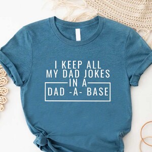 May include: A teal blue t-shirt with white text that reads "I keep all my dad jokes in a dad-a-base".